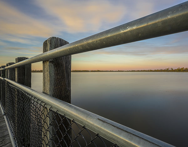 A serene waterscape featuring a wooden pier with a metal railing and chain-link fence, reflecting a colorful sky over calm water.