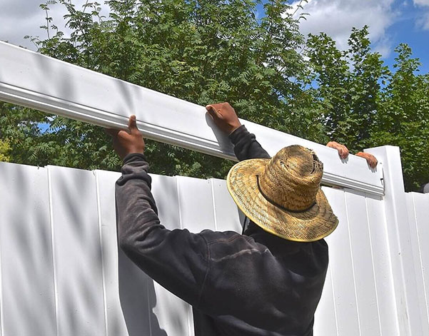 A person in a straw hat installs a white fence panel under a blue sky, while another hand assists from behind.
