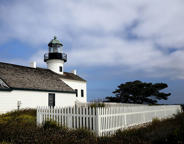 White lighthouse with green lantern, surrounded by a white picket fence and coastal landscape under a cloudy sky.